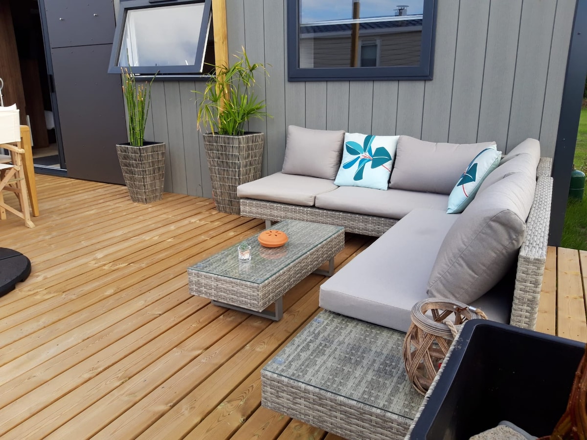 A semi-covered terrace features a modern seating area with a sectional sofa and a low coffee table. Two decorative planters with green plants add a touch of nature. Cushions in shades of blue and white enhance the inviting space, with a warm wooden deck completing the look.