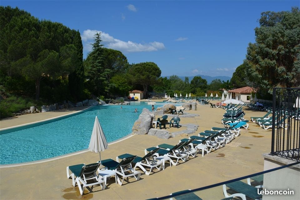 A spacious pool area is surrounded by lush greenery, with a large swimming pool featuring a gentle curve. Numerous lounge chairs, some with umbrellas, are arranged neatly along the poolside, creating an inviting space for relaxation. A few guests can be seen enjoying the water.