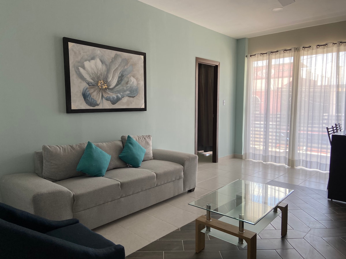 A living area features a light-colored sofa adorned with turquoise cushions, complemented by a glass coffee table. Natural light filters through sheer curtains, illuminating a large floral artwork displayed on the wall. An open doorway leads to additional spaces.
