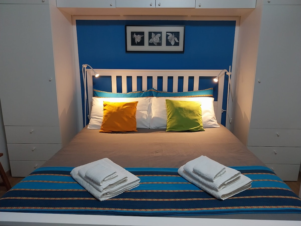 A spacious bed is centered against a vibrant blue wall, featuring decorative pillows in bright yellow and green. Neatly arranged towels are placed at the foot of the bed. Wardrobe storage is available on either side, providing an organized and comfortable layout.