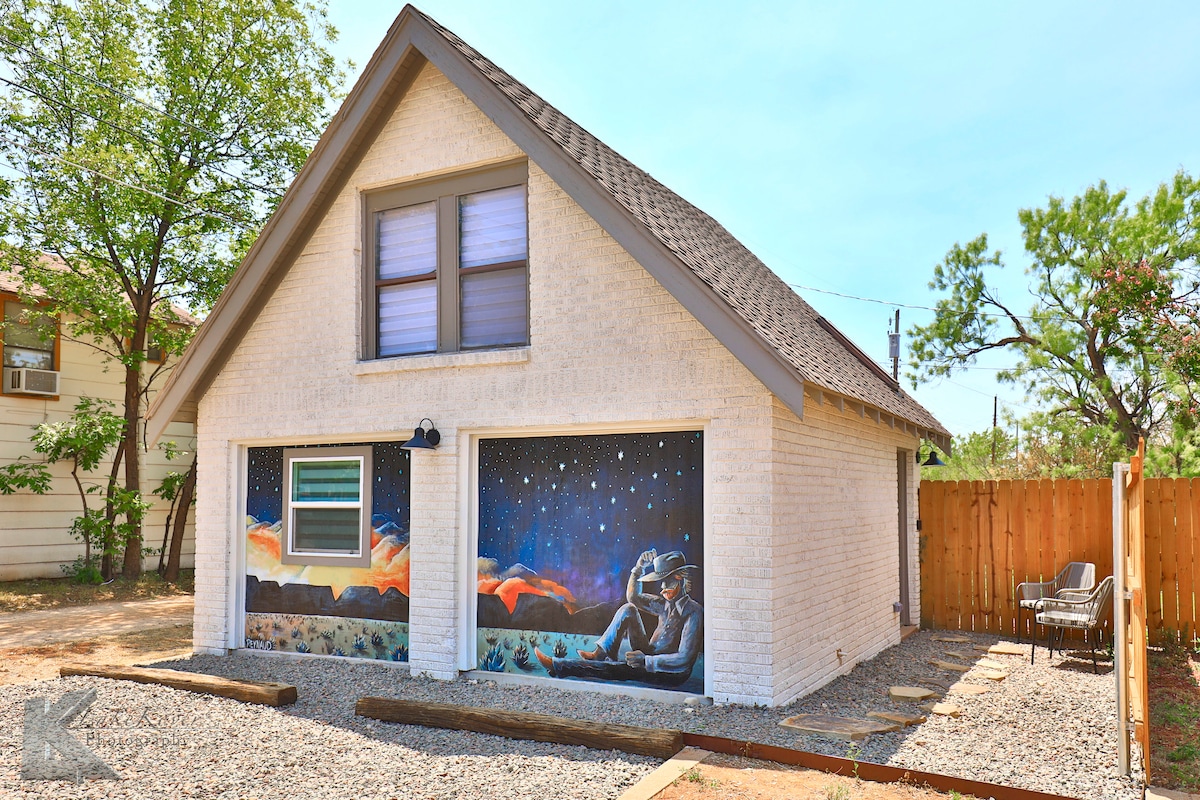 An artistic facade features a mural depicting a peaceful outer space scene, complemented by a window above. The exterior is adorned with light-colored brick and surrounded by gravel, with a small seating area visible on the side, under the shade of nearby trees.
