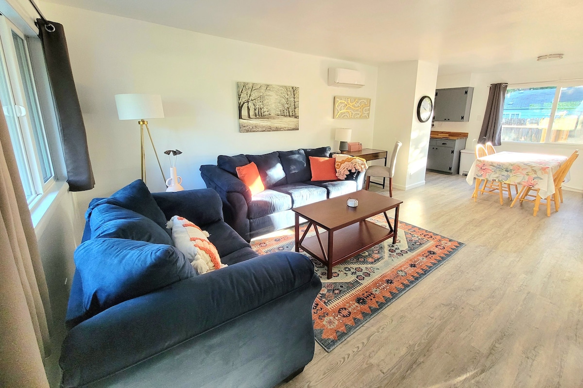 The living room features a spacious seating area with a dark blue couch, accented by decorative pillows. A wooden coffee table is centered on a colorful area rug. Natural light enters through large windows, with a dining area equipped with a wooden table and chairs visible in the background.