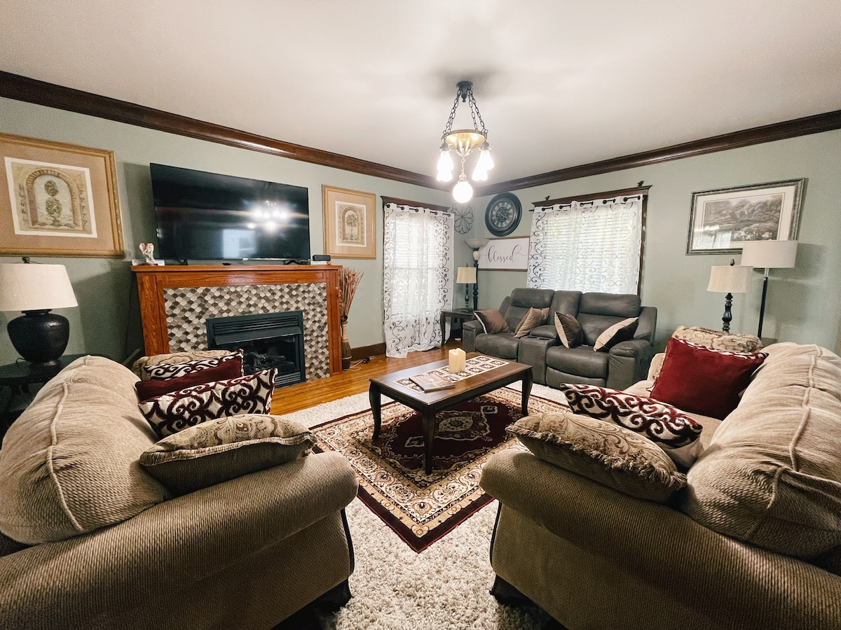 A cozy living area features a decorative fireplace framed by two comfortable couches adorned with plush pillows. A large television is mounted above the fireplace, while gentle light is provided by a central fixture and two side lamps. Soft, neutral tones create a relaxing environment.