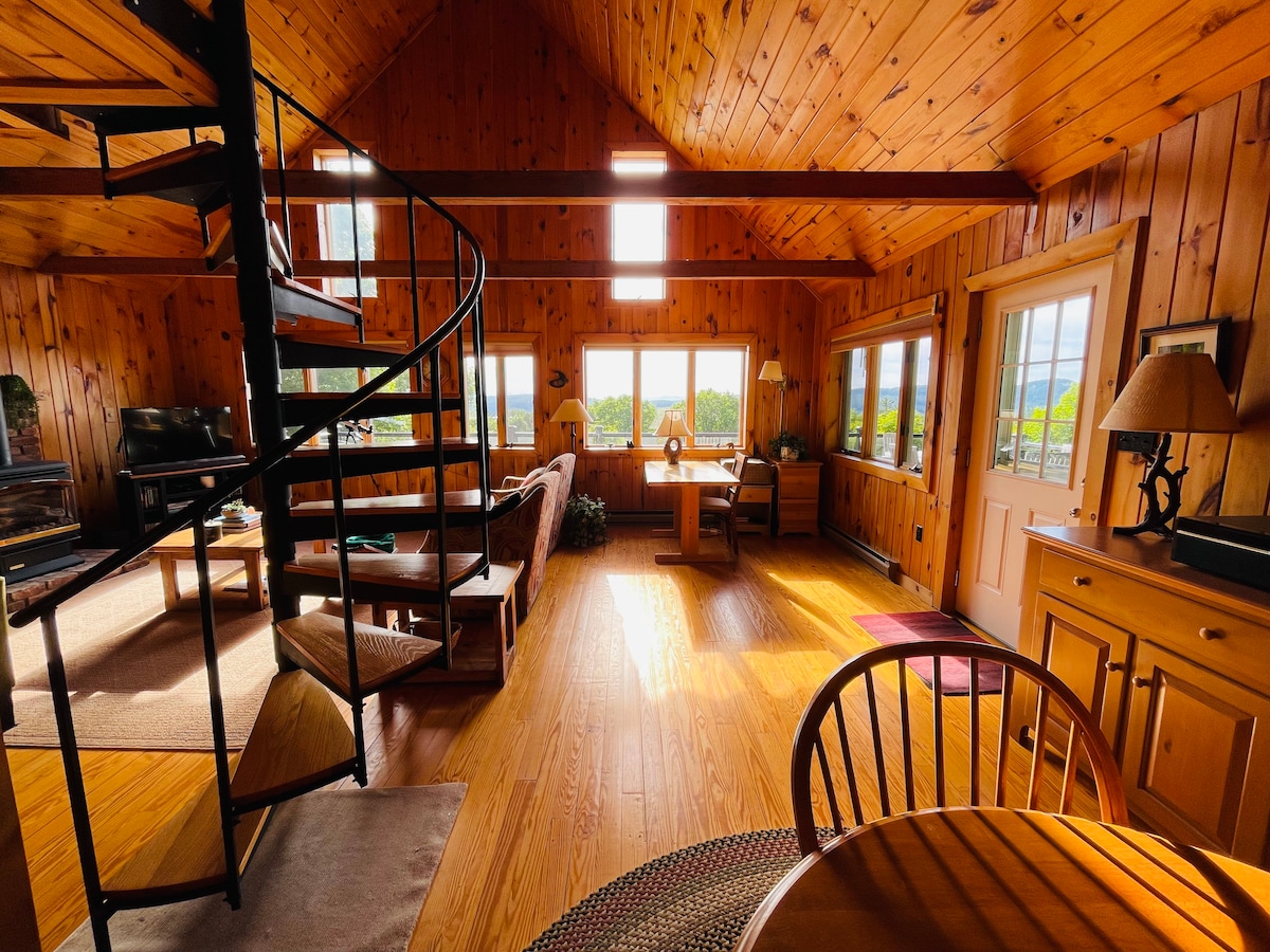 An open living area is highlighted by warm wooden walls and a spiral staircase. Natural light filters through multiple windows, illuminating seating areas and a wooden dining table. A cozy corner workspace is positioned alongside the entrance, emphasizing a relaxed atmosphere.