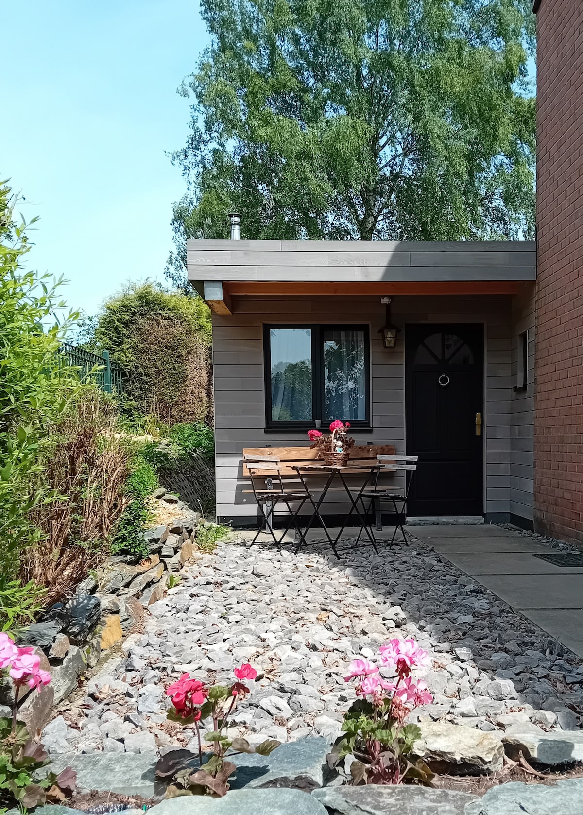 An inviting entrance features a small wooden table set with flowers, surrounded by two folding chairs. A pathway lined with stones leads to a dark front door, framed by greenery and colorful blooming plants.