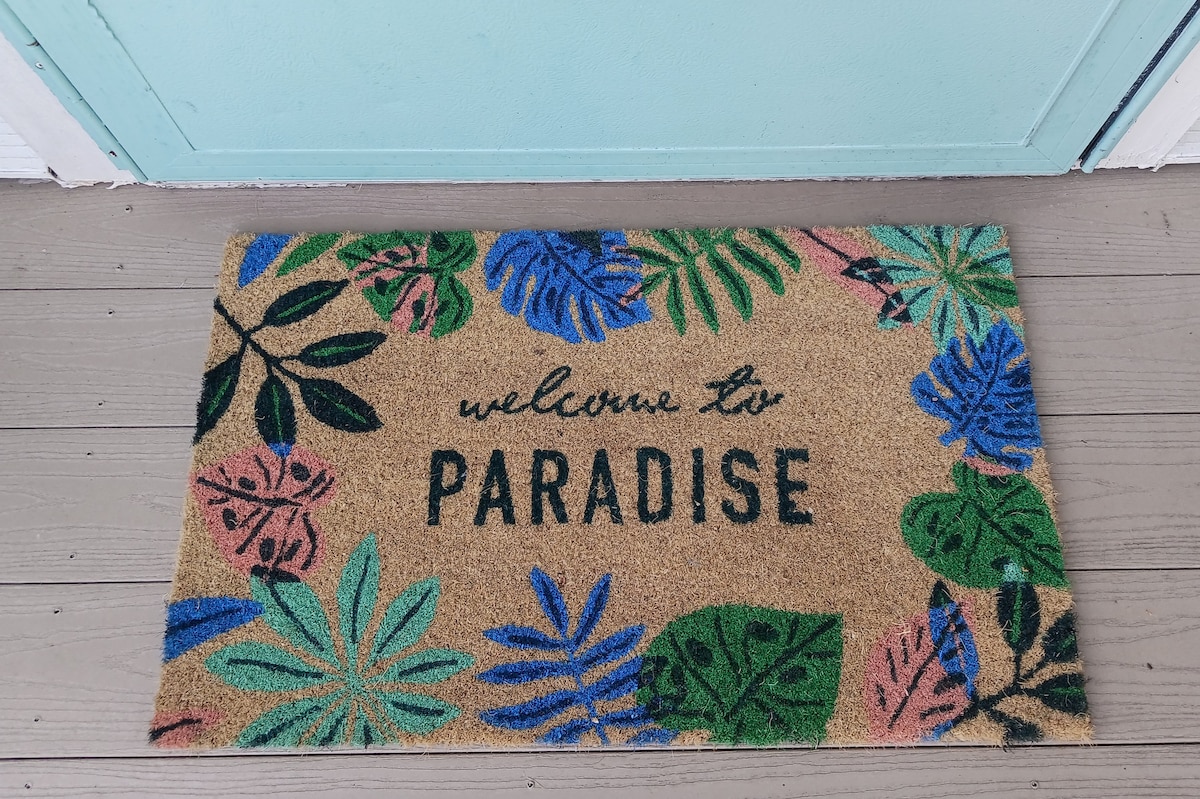 A welcome mat featuring the phrase 'welcome to PARADISE' is displayed at the entrance. The mat is adorned with vibrant tropical leaves in green and pink hues, resting on a light wooden floor in front of a teal door.