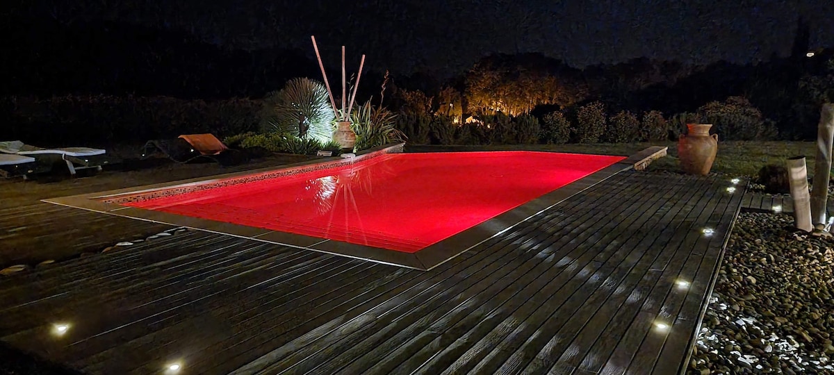A tranquil pool area is illuminated by soft red lighting, creating a serene atmosphere. Surrounding greenery is subtly highlighted, while wooden decking leads around the pool. Comfortable lounge chairs are positioned nearby, and decorative planters add a natural touch to the setting.