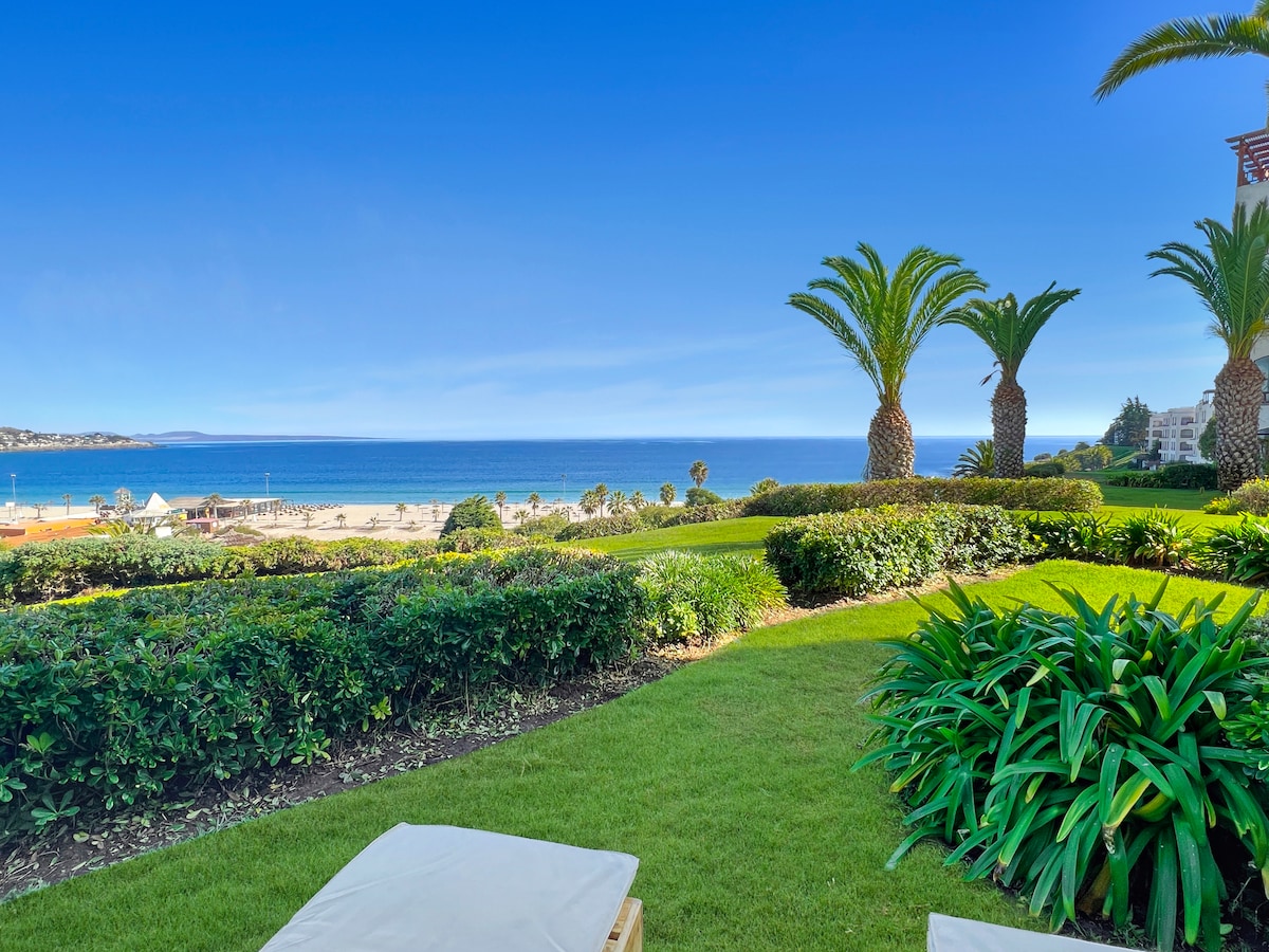 A lush garden area is visible, featuring well-maintained grass and tropical palm trees. The backdrop displays a stunning ocean view, with the beach just beyond the greenery. Clear blue skies enhance the serene atmosphere of the outdoor space.