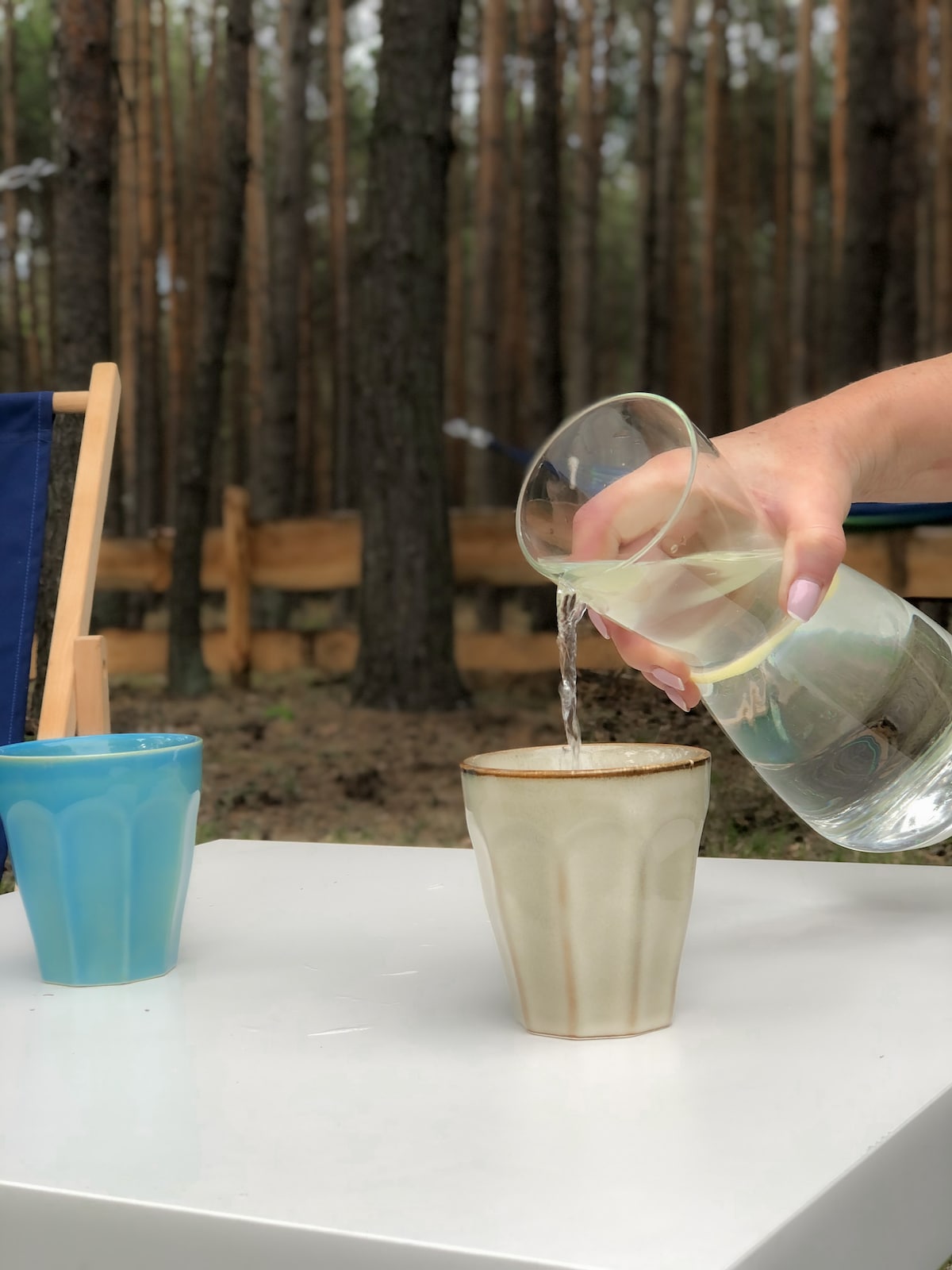 A clear glass is being poured into a decorative cup, with a blue cup beside it. The background features tall trees, creating a serene outdoor setting. A simple white table is visible, enhancing the scene's calm atmosphere.