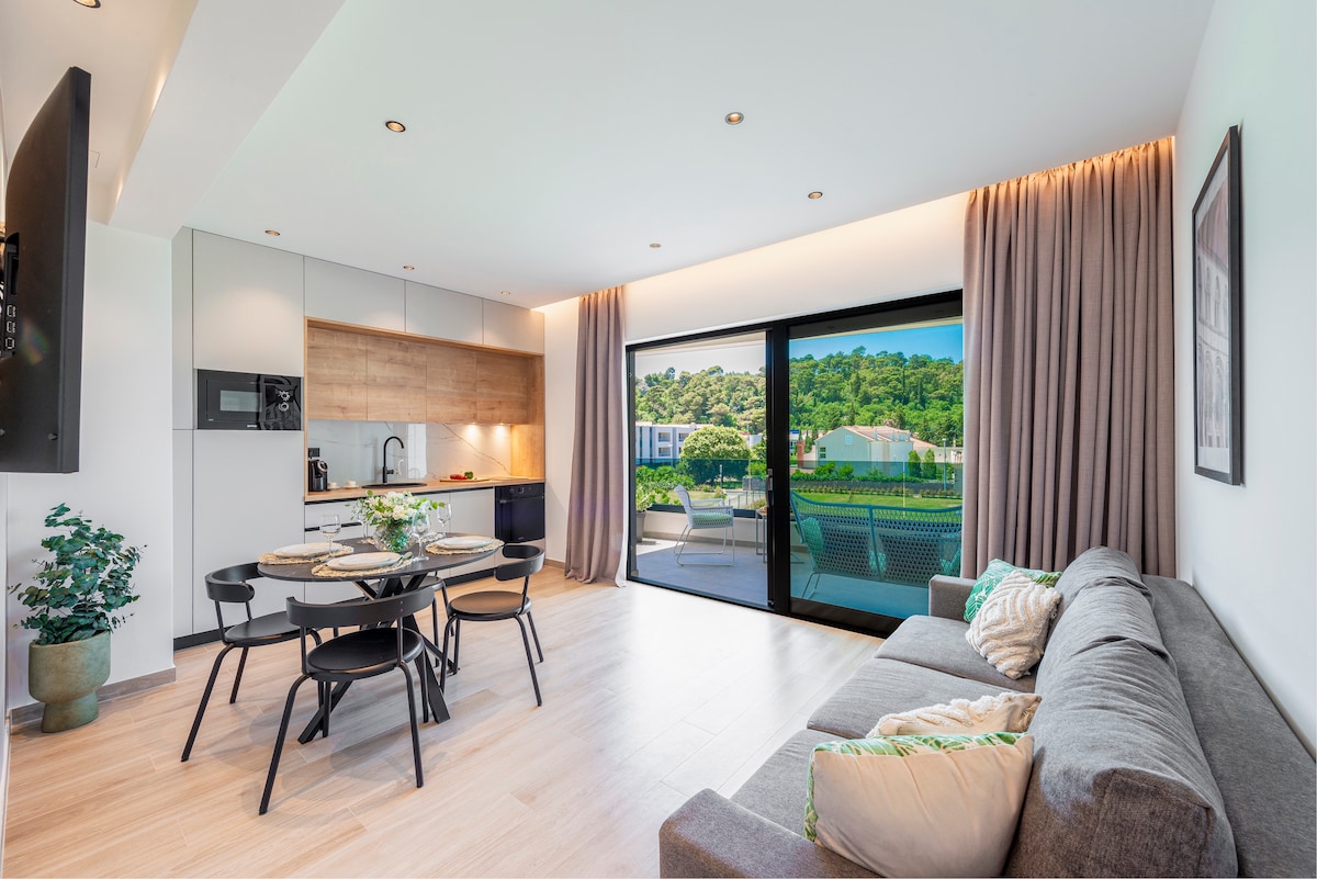 A modern living space features a sleek kitchen area with light wood cabinetry and stainless-steel appliances. A dining table with four black chairs is set for a meal. Large glass doors open to a balcony, inviting natural light and views of greenery into the room.