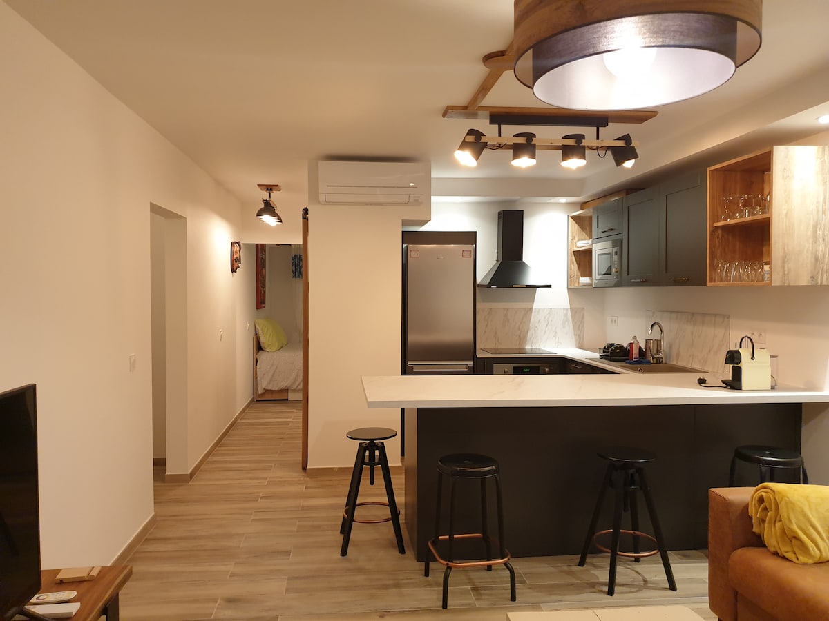 A modern kitchen area features a central island with bar stools, complemented by sleek cabinetry and stainless-steel appliances. Pendant lighting illuminates the space, while warm wood accents add a touch of comfort. A glimpse of the adjoining living area is visible in the background.