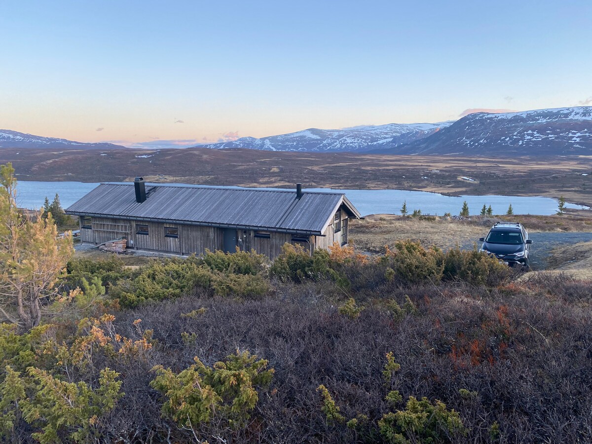 A rustic cabin is nestled among the rugged landscape, with a vast view of mountains and water beyond. The structure is framed by shrubs, and a vehicle is parked nearby, suggesting easy access to the serene surroundings.