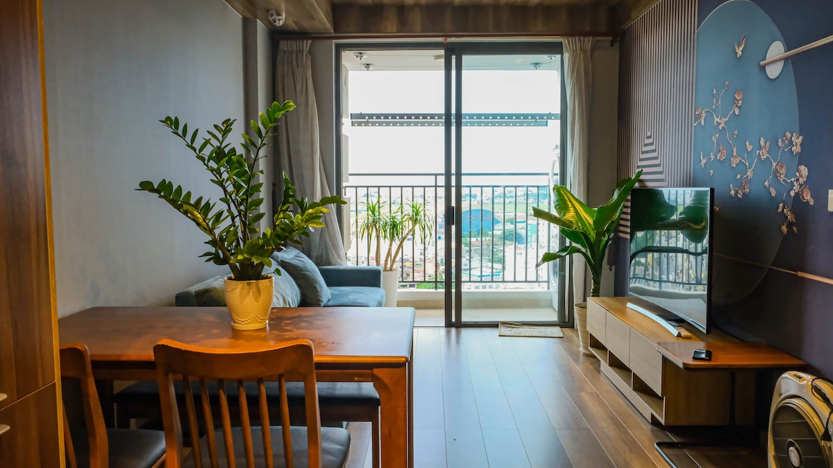 The living room features a sleek wooden dining table accompanied by wooden chairs. A plush sofa is positioned near large glass doors, leading to a balcony with a view. Green plants enhance the space, alongside a modern TV set on a wooden console.