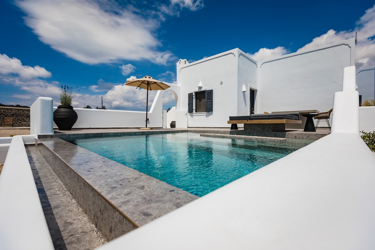 A refreshing pool area is framed by sleek stone edges, with clear blue water reflecting the sky. A sun umbrella offers shade alongside a wooden seating arrangement, while the whitewashed building contrasts against the vibrant outdoor space, creating a serene atmosphere.