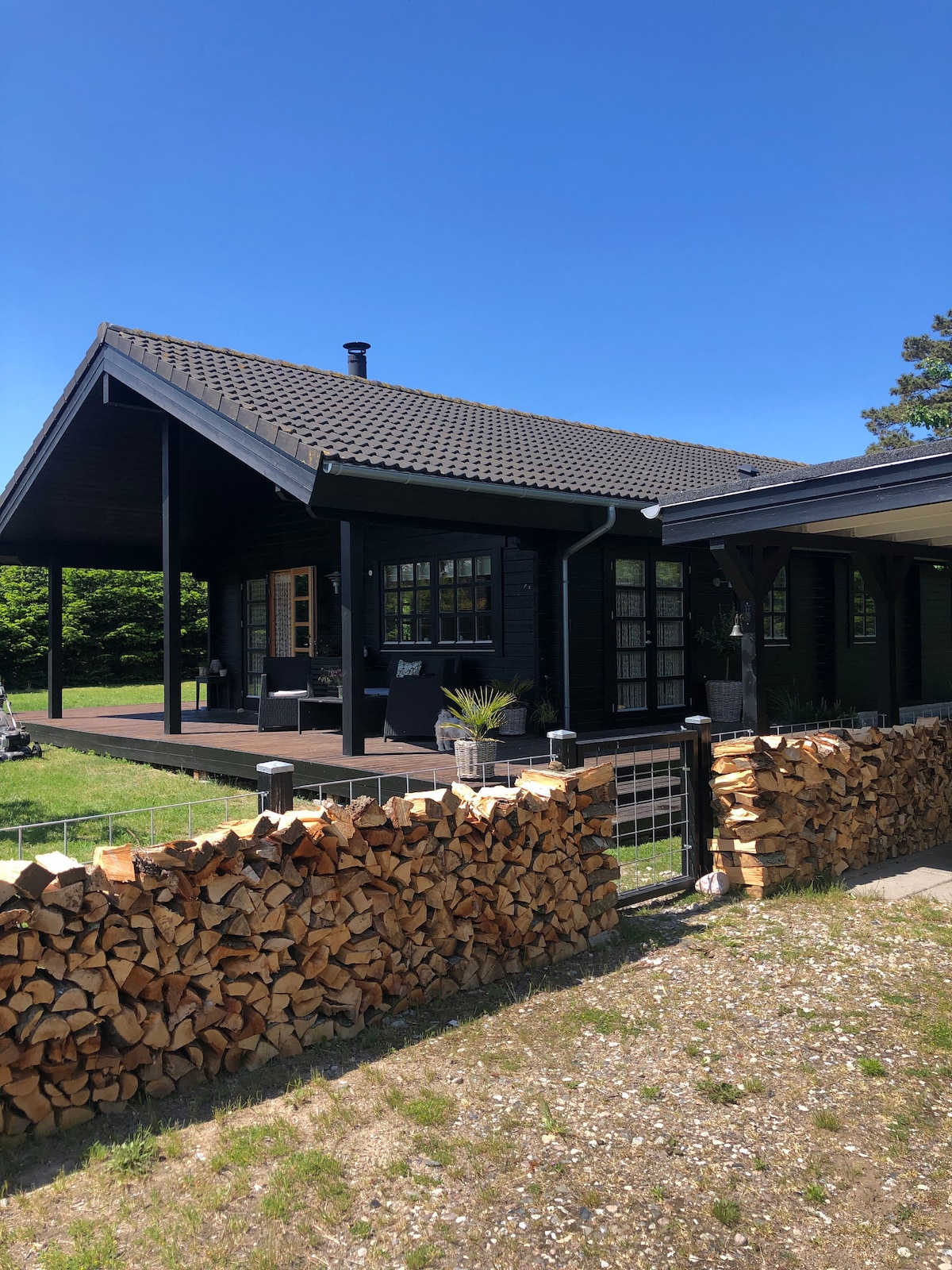 The exterior of a black wooden house is showcased, featuring a sloped roof and bright blue sky above. A porch area is visible with outdoor seating. A neatly stacked woodpile borders the property, complementing the natural surroundings.
