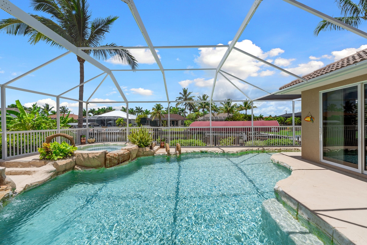 The zero-entry pool features a gentle slope, surrounded by palm trees and expansive screened-in lanai. Sunlight reflects off the sparkling water, while views of lush tropical landscaping create a serene atmosphere. The backdrop showcases a mix of residential rooftops and greenery under a clear blue sky.