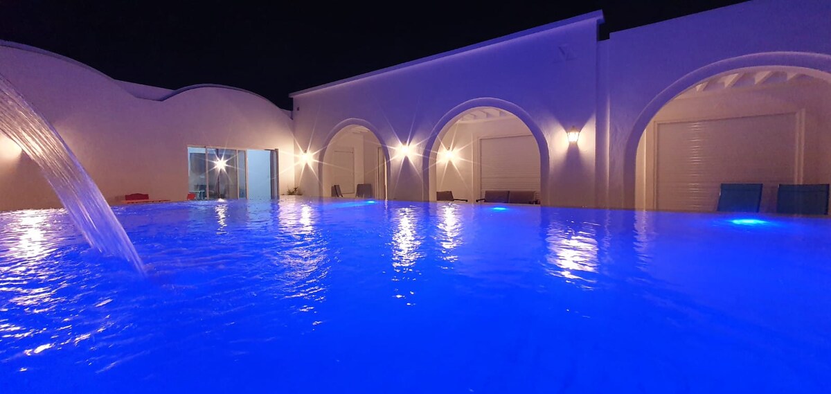 The illuminated pool reflects a soft blue hue under the night sky, featuring a gentle cascade. Arched openings reveal the surrounding structure, enhancing the ambiance with warm lighting. Comfortable loungers are positioned nearby, inviting relaxation in a serene environment.
