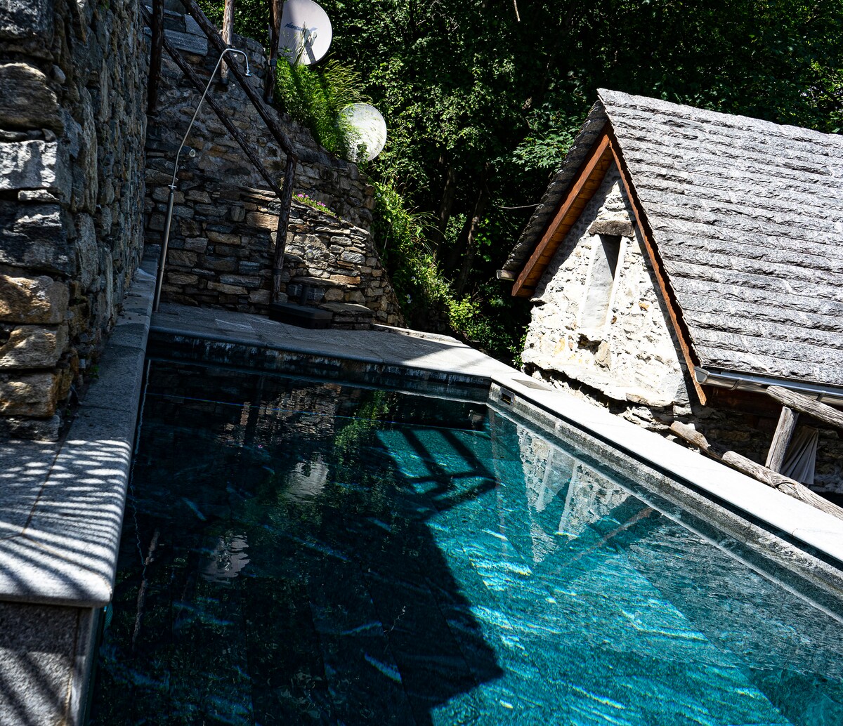 A tranquil swimming pool features clear turquoise water, surrounded by stone walls and lush greenery. A rustic structure with a sloped roof is partially visible, reflecting natural sunlight. The atmosphere is calm and inviting, showcasing a harmonious blend of nature and architecture.