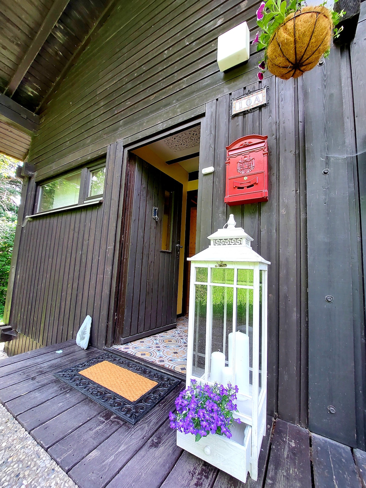 The entrance features a dark wooden facade, complemented by a decorative lantern in white and a vibrant purple flower pot. A welcoming doormat and a vintage-style mailbox complete the inviting entryway.