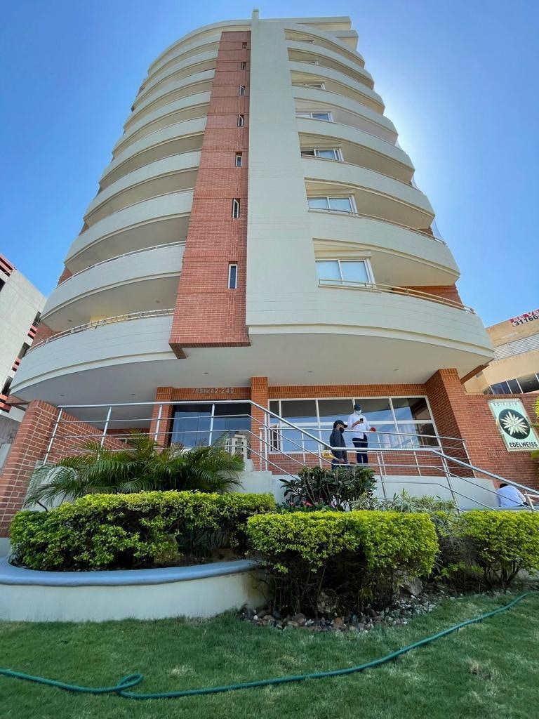 A modern multi-story building features a sleek brick exterior and large windows. A landscaped area with green shrubs and grass surrounds the entrance, which is accessible via a gentle ramp. Natural light illuminates the sidewalk, enhancing the building's contemporary design.