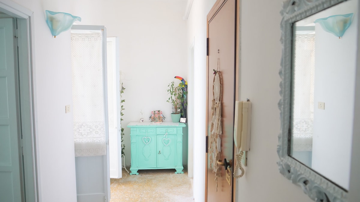 A bright hallway is shown, featuring light-colored walls and an inviting atmosphere. A vintage mint-green cabinet adds a pop of color, while natural light pours in through an open doorway. A lovely mirror is mounted on the wall, enhancing the space.