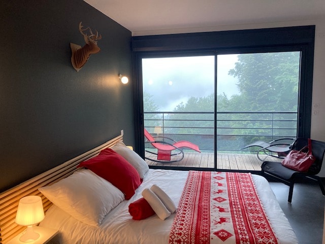 A cozy bedroom features a spacious bed adorned with a patterned blanket and plush pillows. Large sliding glass doors open to a private balcony, offering views of a misty landscape. A minimalist design is complemented by a modern chair and decorative wall art.