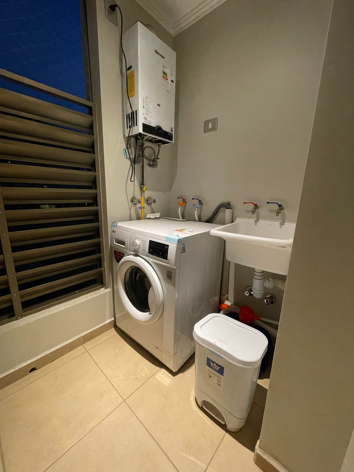 A functional laundry area is presented, featuring a washing machine beside a utility sink. The environment is illuminated softly, highlighting the organized plumbing fixtures. A trash bin is located on the floor, and windows with slatted coverings allow natural light while maintaining privacy.