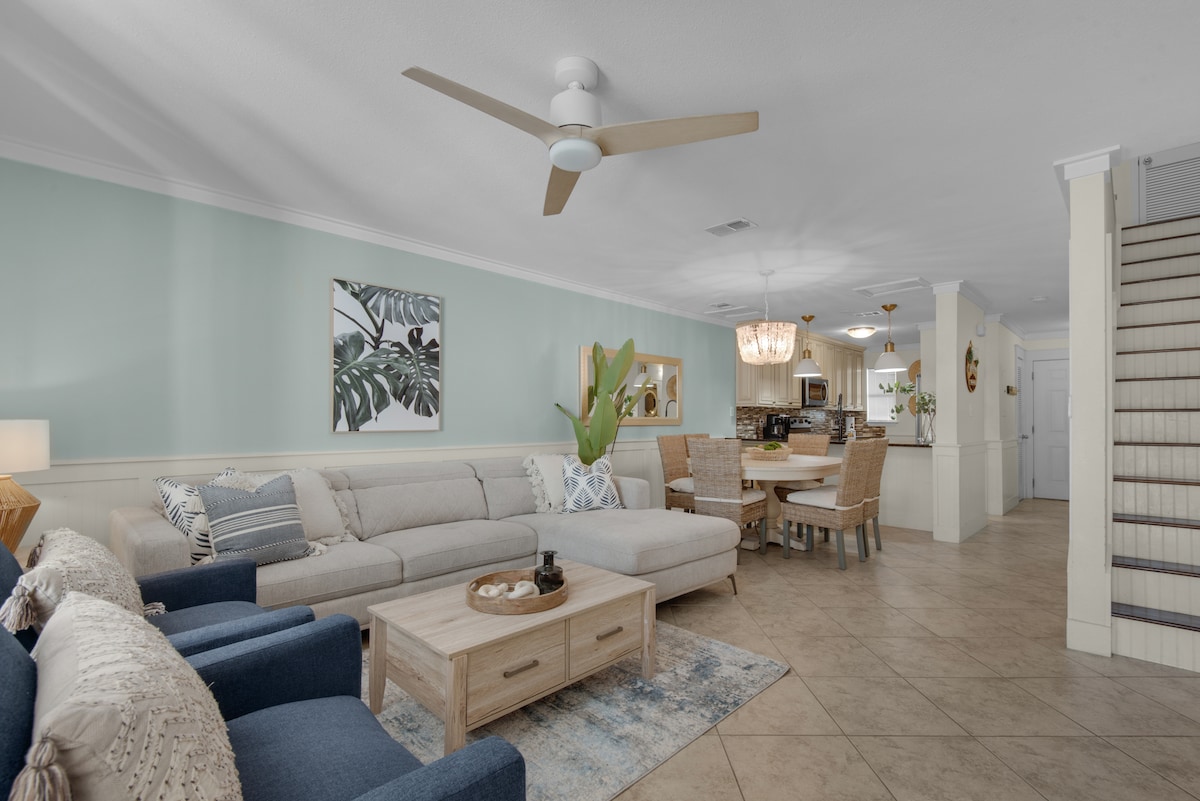 A spacious living area features a light-colored sectional sofa and two blue armchairs, complemented by a wooden coffee table. The dining area includes a round table with seating for six. Soft green walls and decorative plants enhance the inviting atmosphere.