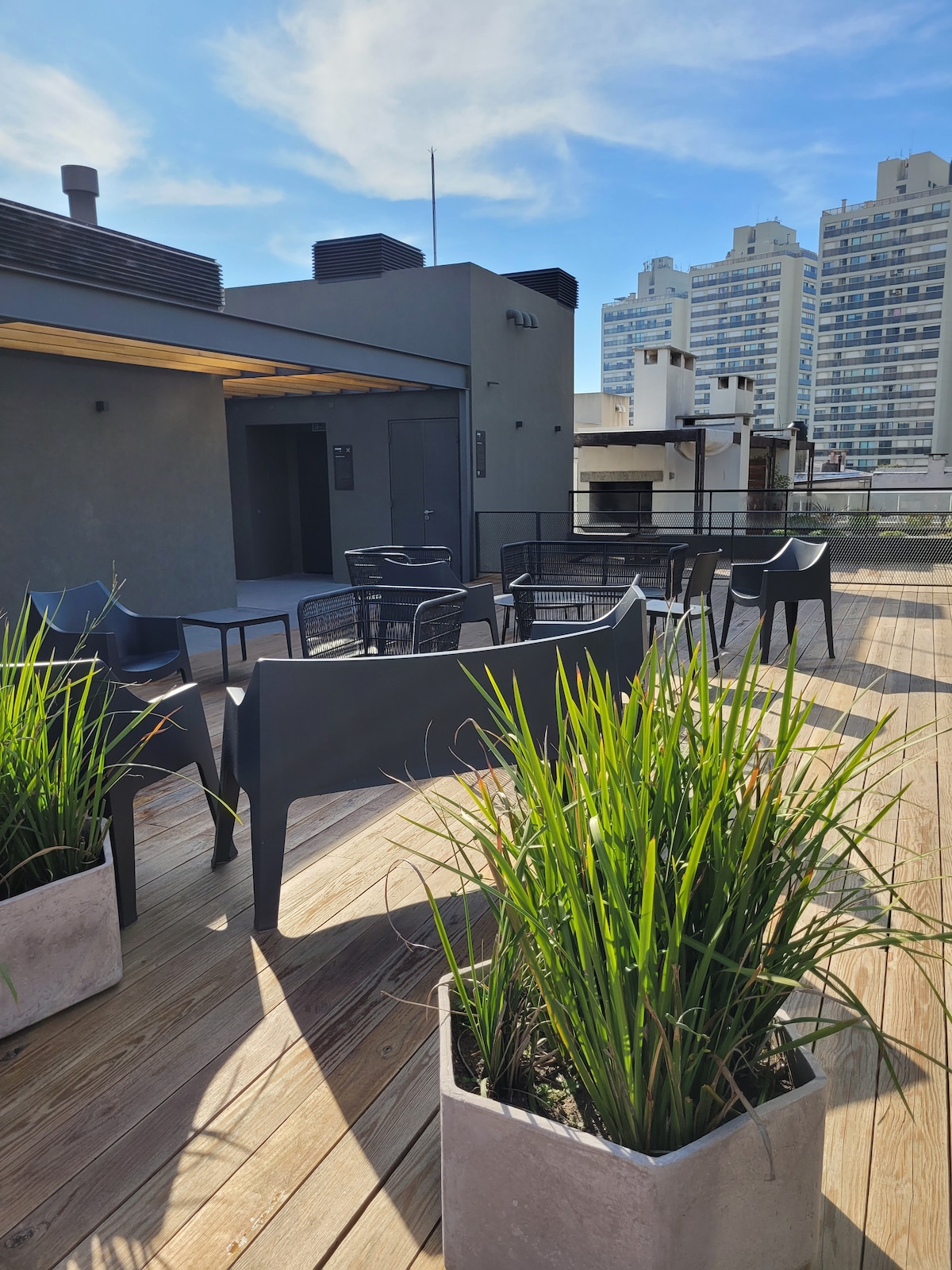 An outdoor terrace features several black chairs and a large table, surrounded by potted green plants. The wooden deck extends across the space, with a backdrop of tall buildings under a clear blue sky.