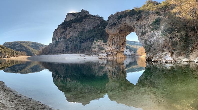 A natural rock arch is reflected in the calm waters of a river, surrounded by lush greenery. The landscape features gently sloping hills in the background, with a sandy bank leading to the water's edge. The scene captures a serene and tranquil environment.