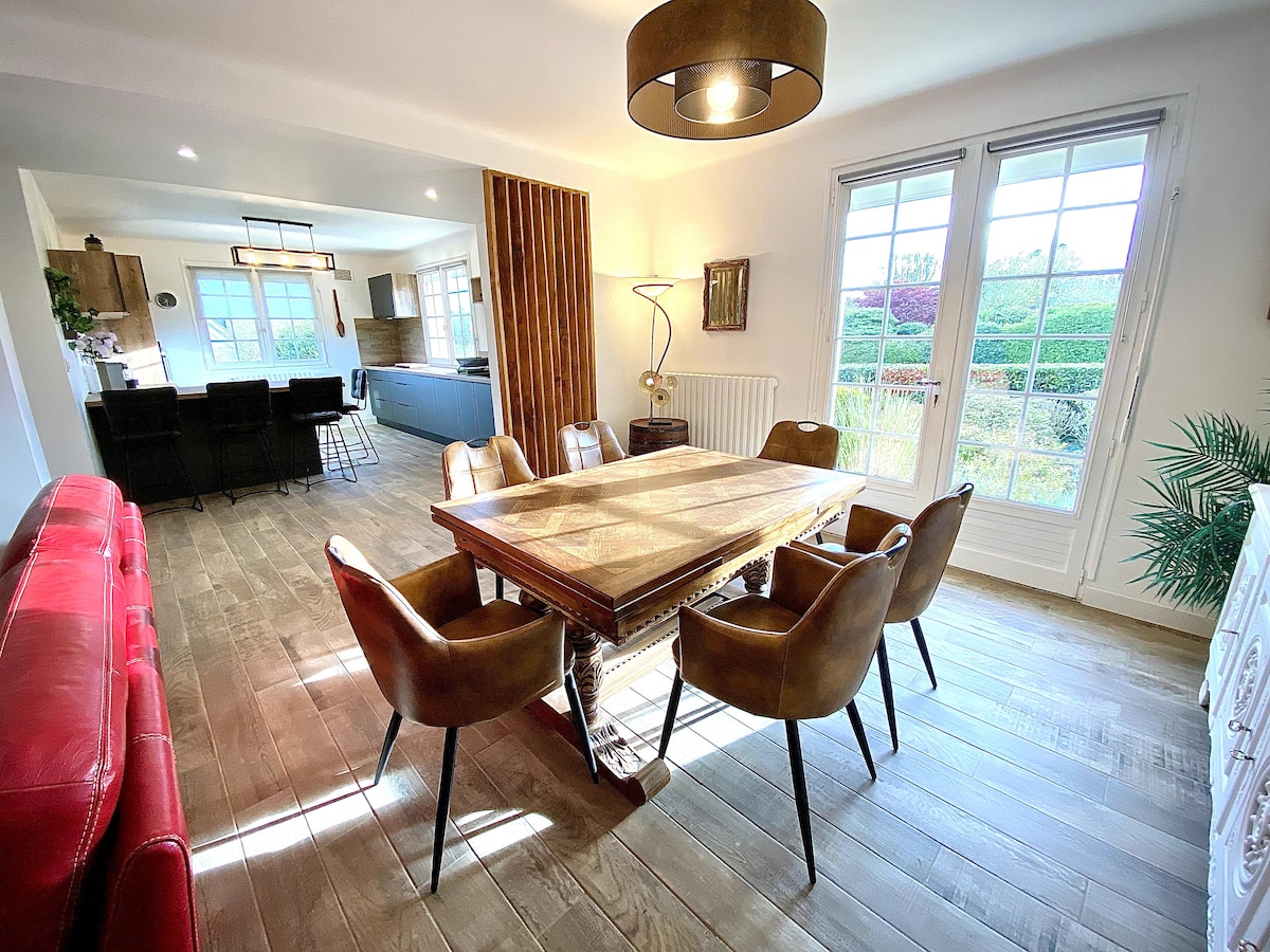 A dining area features a rustic wooden table surrounded by six plush, brown chairs. Natural light floods through large French doors, illuminating the space and offering views of a green landscape. A modern pendant light hangs above the table, enhancing the ambiance.