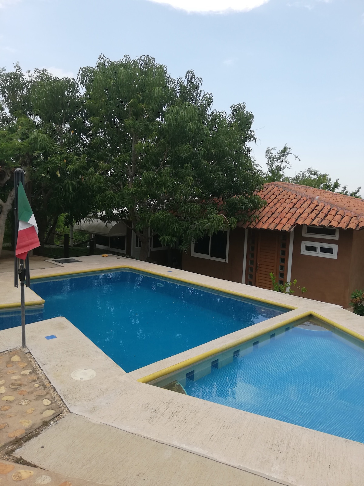 A shared pool area is surrounded by lush greenery and includes two distinct swimming sections. A flag represents the local culture nearby. The cozy architecture of the adjacent building harmonizes with the natural landscape.