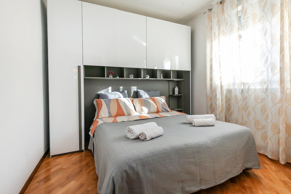 A spacious bed is adorned with neutral-toned linens, accompanied by neatly arranged towels. Decorative pillows add a touch of style. Natural light filters through sheer curtains, illuminating the room, which features a minimalistic wardrobe and shelving unit against a light-colored wall.