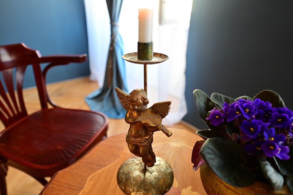 An ornate candle holder featuring an angel figurine is positioned on a wooden table. A lit candle rests atop the holder, surrounded by a fresh arrangement of violet flowers in a green pot. A wooden chair is visible in the background, adding to the inviting ambiance.