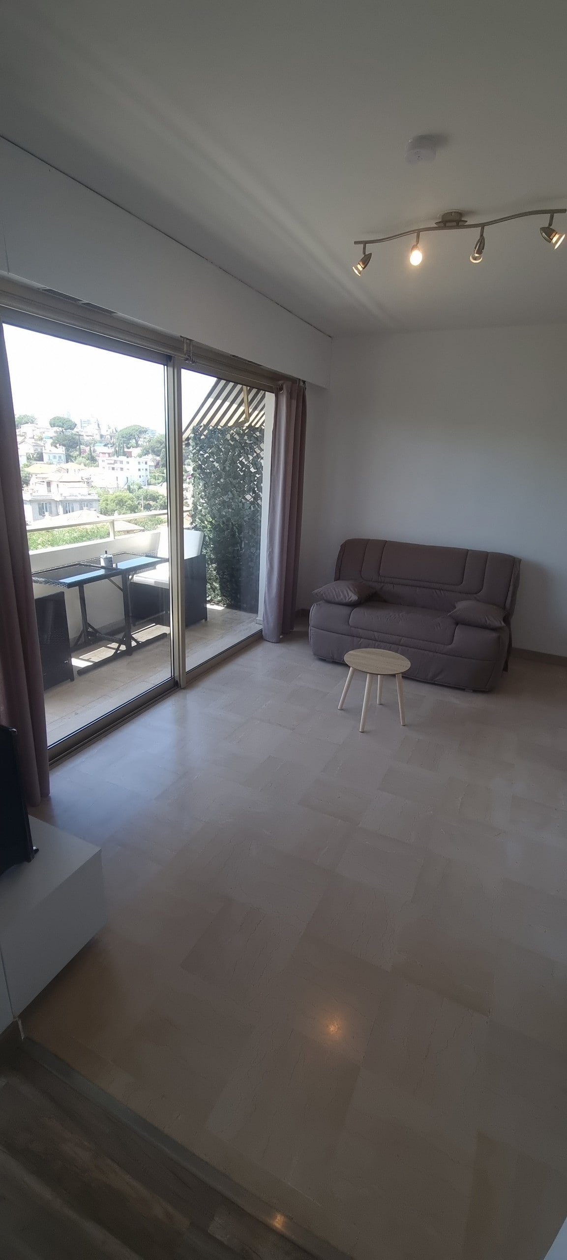 A spacious living area features a convertible sofa and a small wooden stool. Large glass doors allow natural light to fill the room and open to a terrace with outdoor seating, offering views of the surroundings.