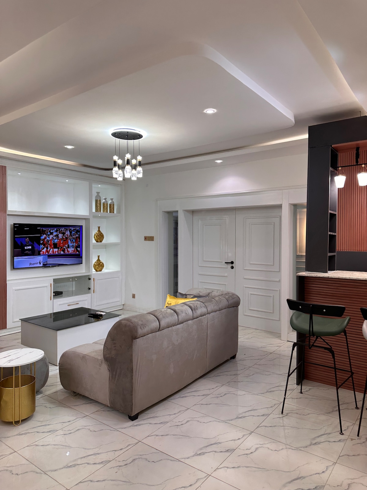 A modern living area features a plush sofa facing a wall-mounted television. Soft lighting is provided by a chandelier, while sleek furniture enhances the space. A marble side table and gold accents add elegance to the serene white and gray color palette.