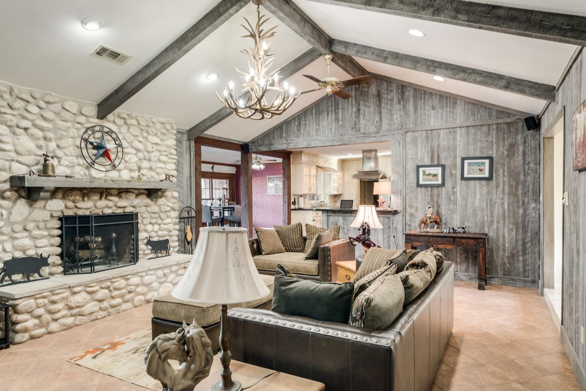 A spacious living area features a high, vaulted ceiling accented by wooden beams. A stone fireplace serves as a focal point, while a leather sofa and comfortable chairs invite relaxation. Soft lighting from a chandelier and table lamps enhances the cozy ambiance.