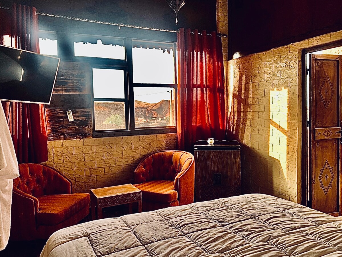 A cozy sleeping area features a spacious bed covered with a light quilt. Two orange armchairs sit beside a small wooden table, and natural light filters through large windows adorned with dark red curtains. A television is mounted on the wall opposite the bed.