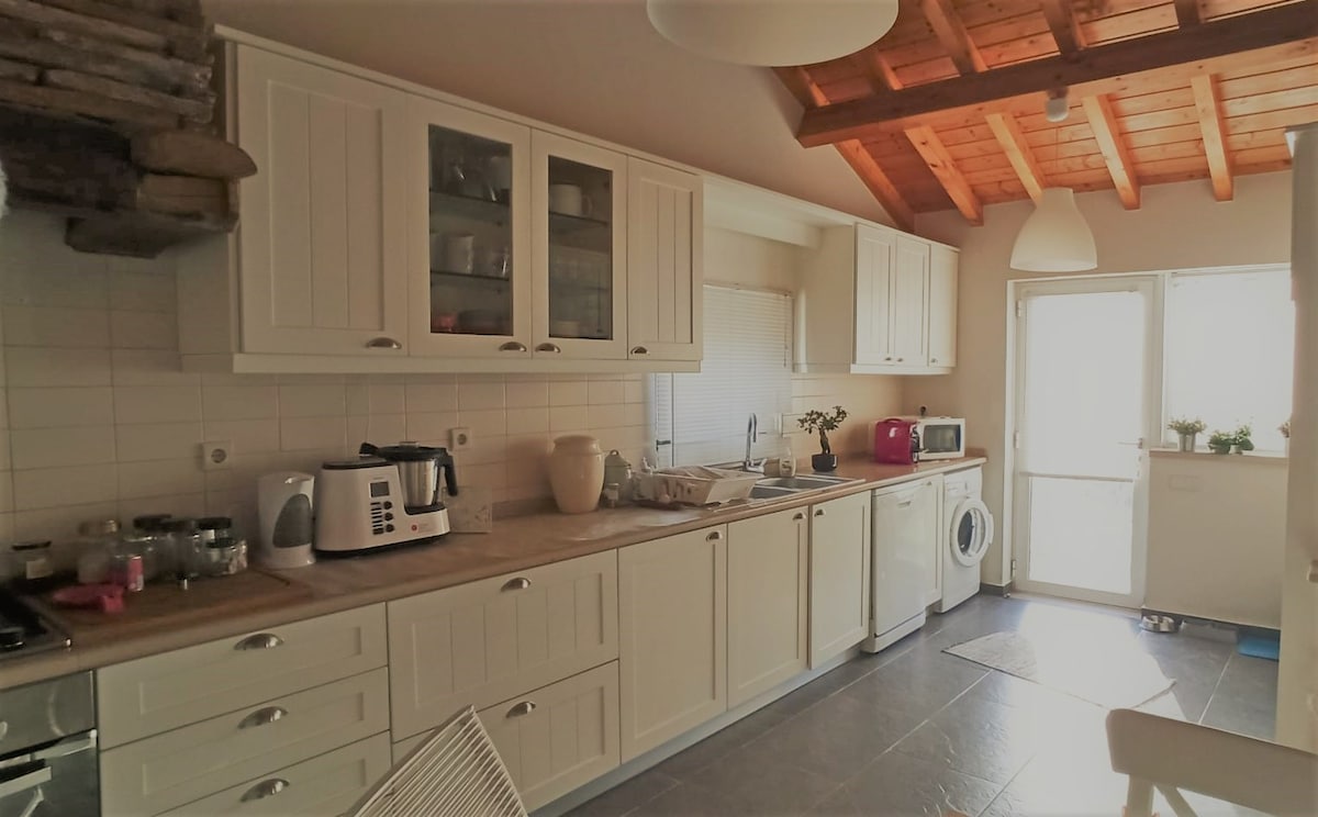 A modern kitchen features white cabinetry with glass accents, providing ample storage. Stainless steel appliances, including a kettle and microwave, are visible. A washer is positioned near the window, which allows natural light to illuminate the space. The ceiling showcases decorative wooden beams.