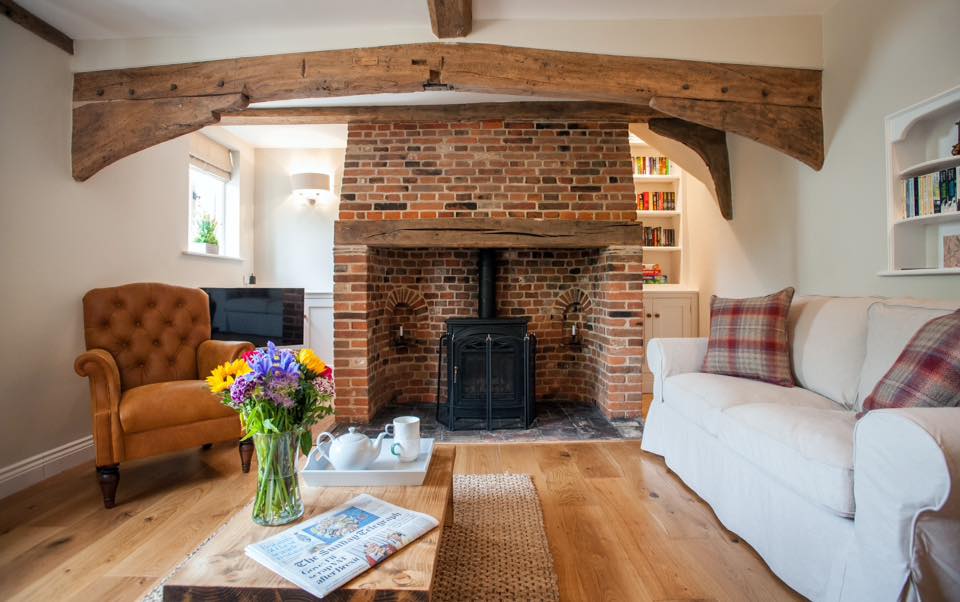 A cozy living area features a striking inglenook fireplace made of exposed brick, with a wood-burning stove at its center. Natural wooden beams span the ceiling, while a comfortable sofa, adorned with decorative pillows, sits alongside an armchair. A wooden coffee table holds a vase of fresh flowers and reading materials.