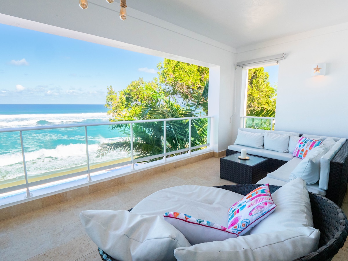 An oceanfront balcony is presented with a view of rolling waves and lush greenery. Outdoor seating includes a cozy sectional sofa adorned with colorful cushions. The tranquil scene invites relaxation, while natural light floods the space through large glass sliding doors.