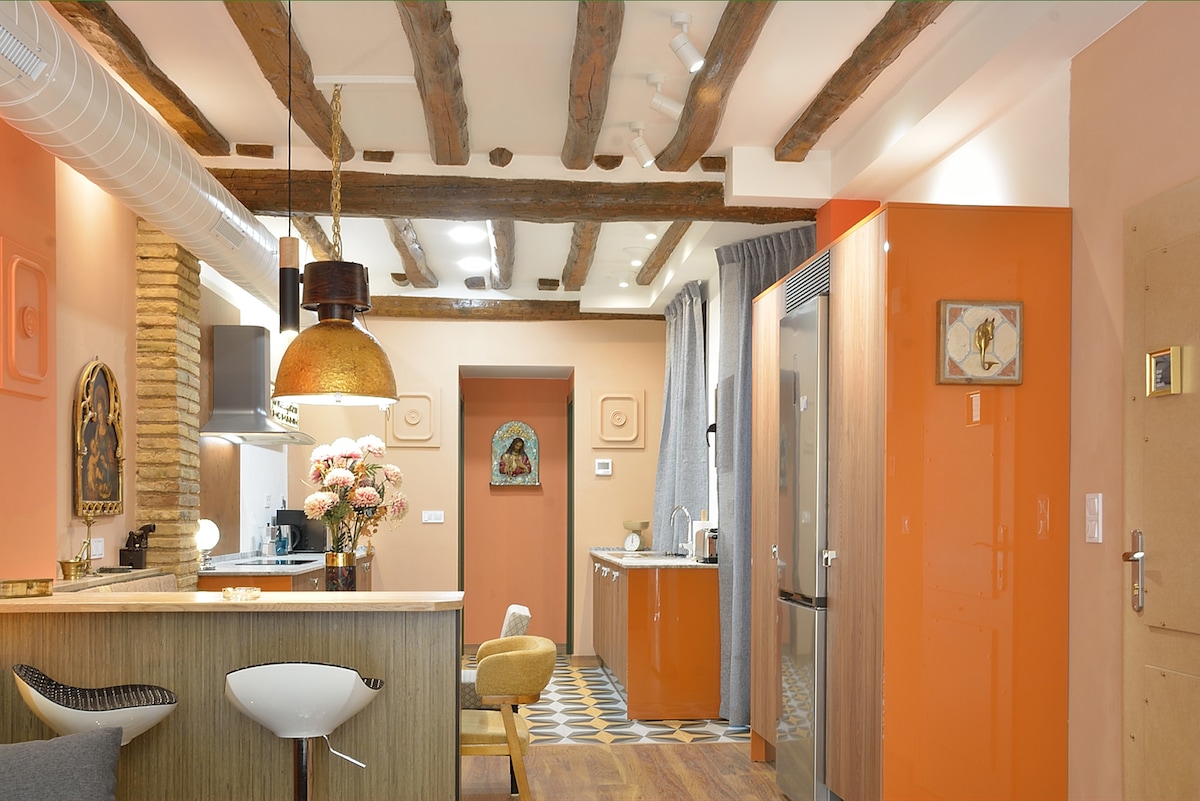 An open living space features rustic wooden beams overhead and a mix of warm orange tones on the walls. A modern kitchen is equipped with essential appliances, while a stylish bar area includes a sleek stool. Decorative objects and flowers add touches of color to the setting.