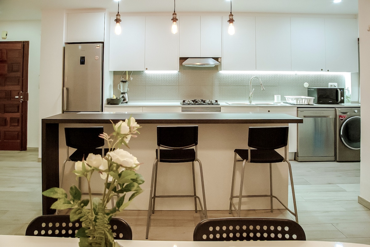 A modern kitchen is displayed with sleek cabinetry and under-cabinet lighting. A central island with high stools is complemented by stainless steel appliances, including a refrigerator and a washing machine. Natural light illuminates the space, enhancing its clean and inviting design.