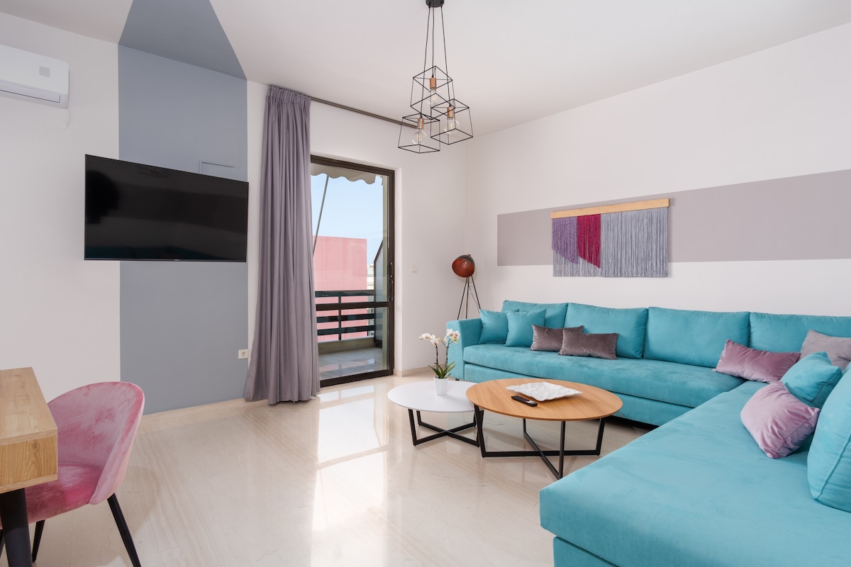 A modern living room showcases a large blue sofa paired with pastel cushions. A flat-screen TV is mounted on the wall, while a round coffee table sits in front. Large sliding doors lead to a balcony, allowing natural light to illuminate the space.