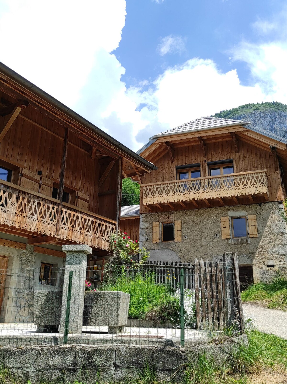 Two charming chalets are set against a mountainous backdrop, showcasing a combination of wood and stone architecture. One chalet features a spacious balcony with decorative wooden railings. Greenery surrounds the stone wall and a gate, enhancing the natural setting.
