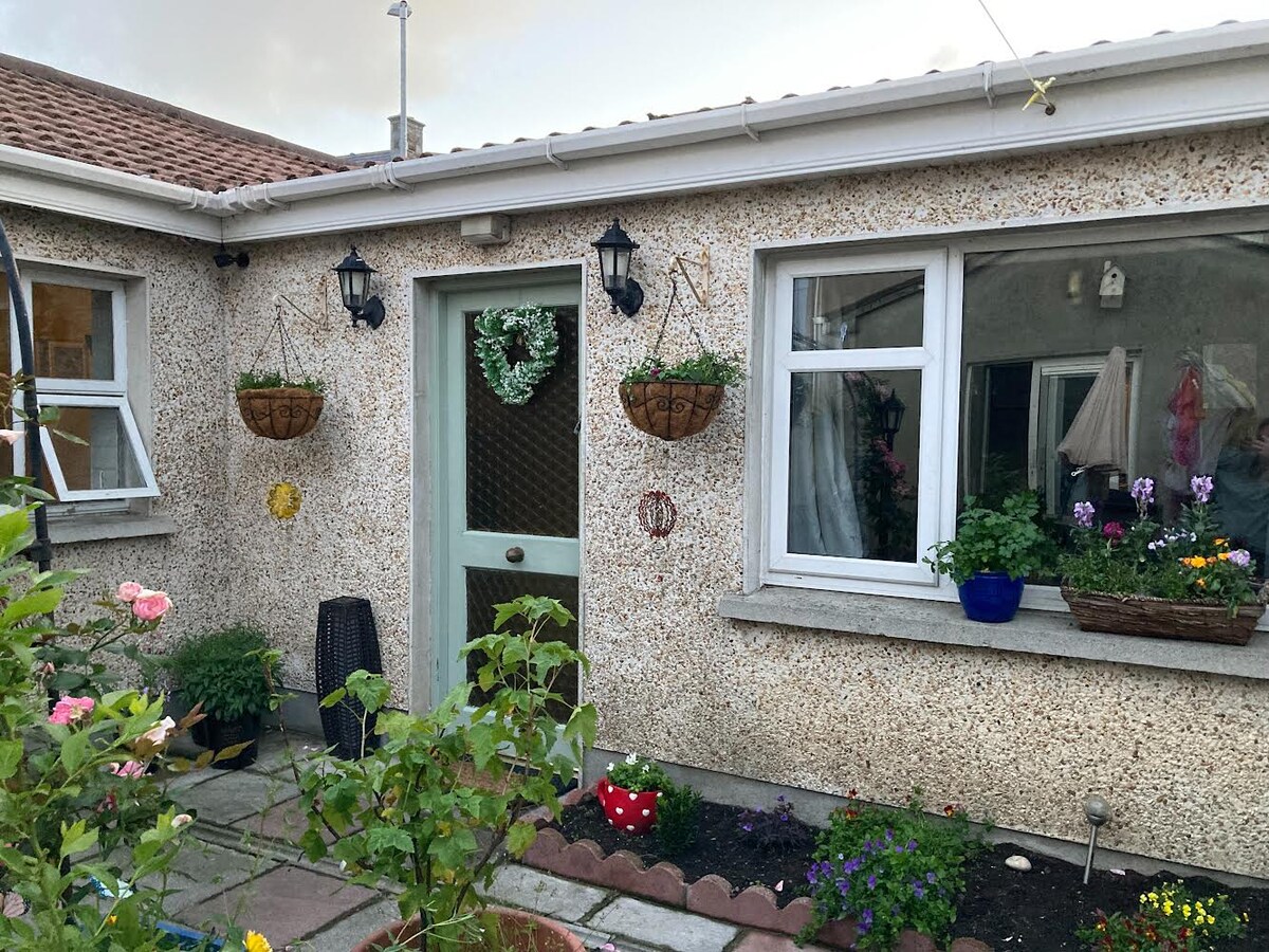 The exterior of the cottage presents a charming facade in a light texture, enhanced by hanging flower baskets and potted plants. A decorative door is flanked by two windows, which feature vibrant greenery and colorful flowers in the nearby garden.