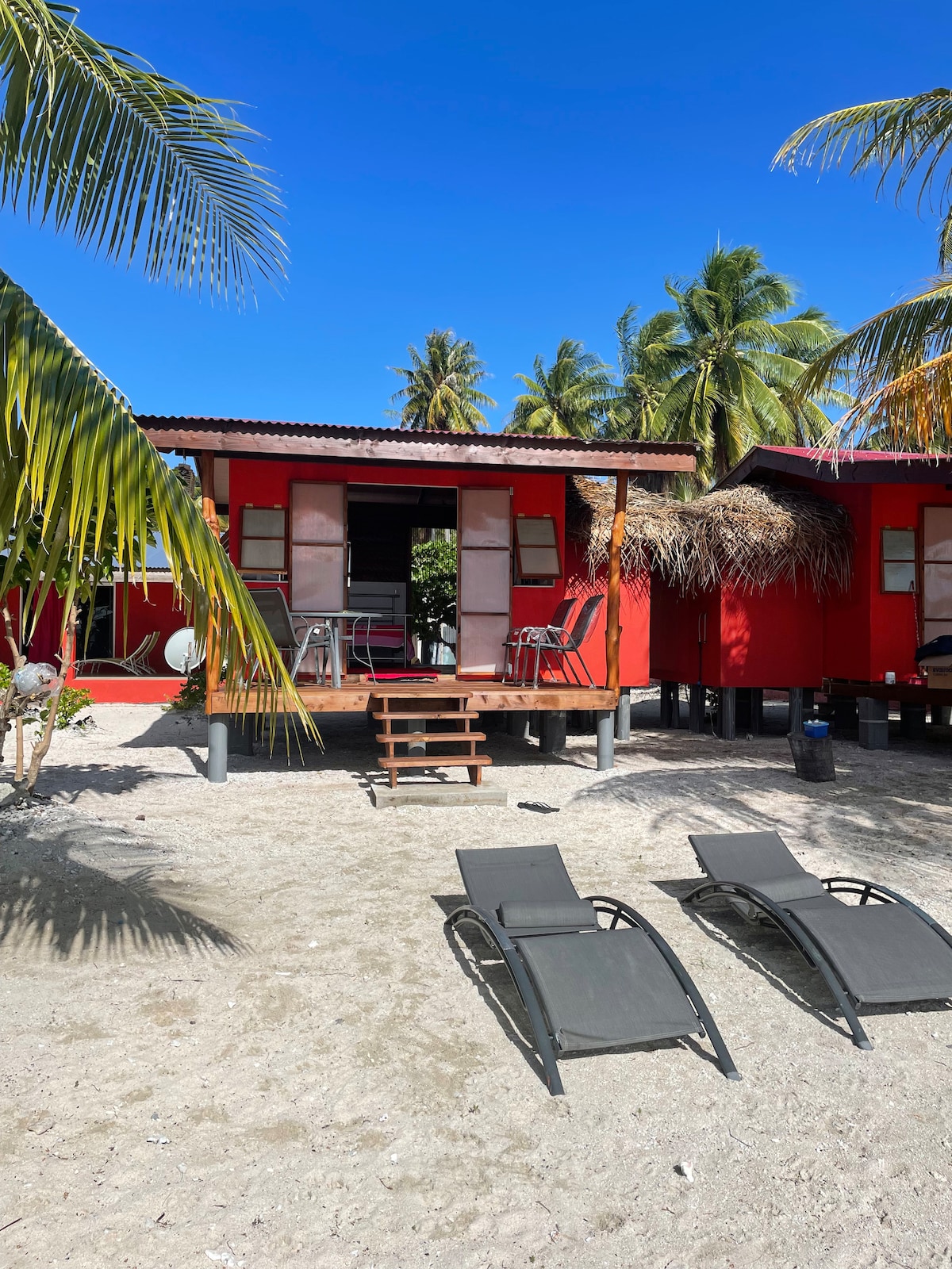The vibrant red bungalow is situated on sandy ground, surrounded by palm trees. A set of steps leads to a shaded deck with outdoor seating, while two lounge chairs are positioned nearby, inviting relaxation in a serene beach setting.