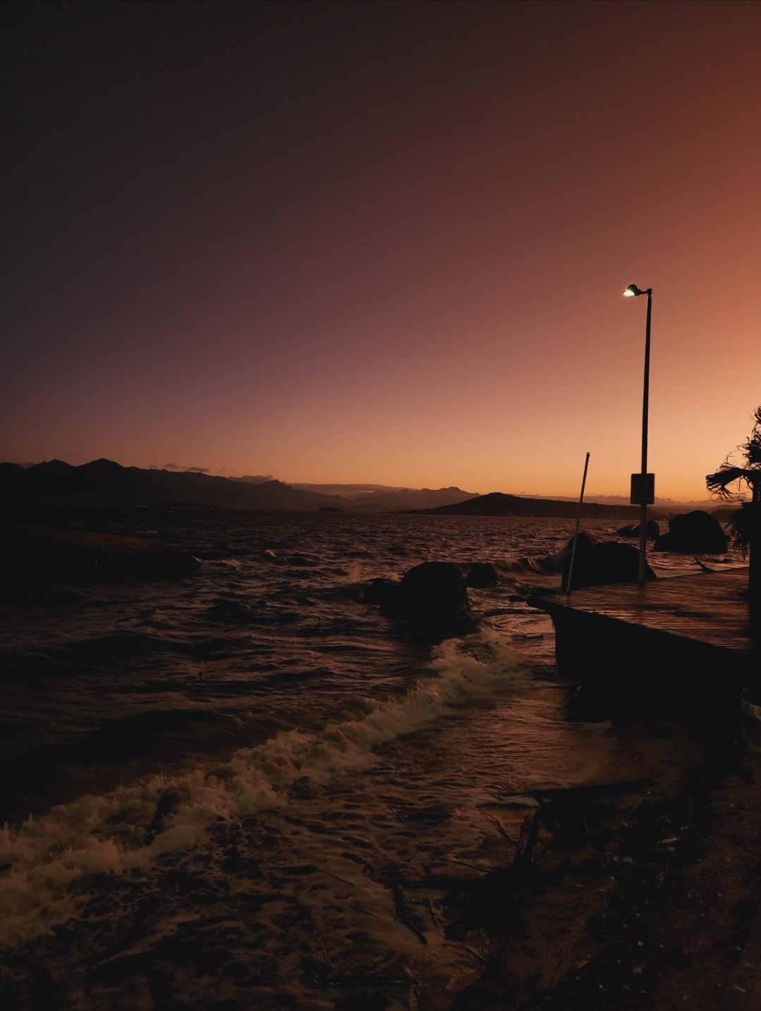A serene coastal view at sunset is depicted, with soft waves gently lapping against rocky shores. The sky transitions from orange to purple, while a solitary lamppost provides illumination. Silhouettes of distant mountains are visible against the horizon, enhancing the peaceful ambiance.