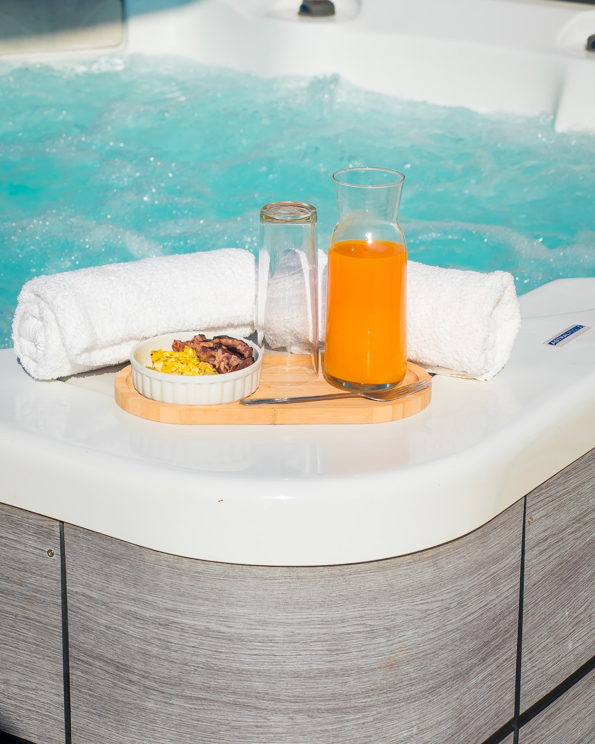 A well-arranged spa corner features a jacuzzi filled with bubbling water. Nearby, a wooden tray holds a colorful drink in a pitcher and a small bowl of food. Soft white towels are neatly placed, contributing to the inviting wellness experience.