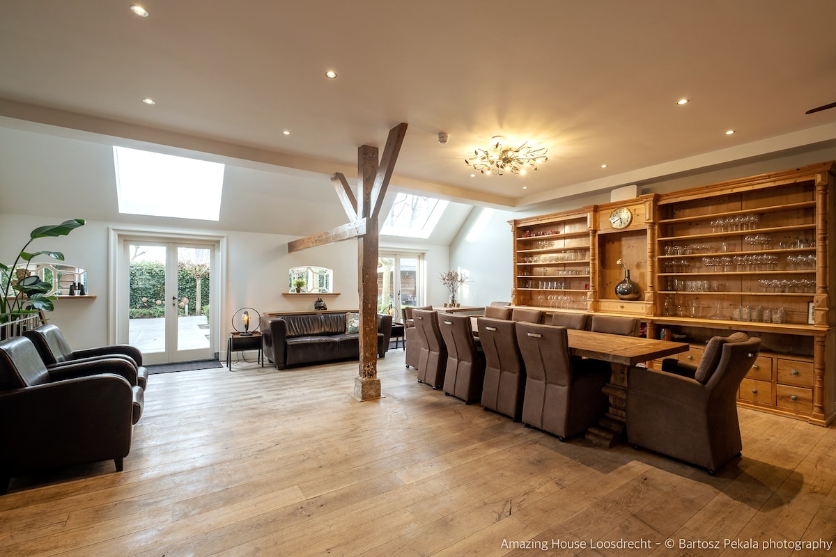 A spacious living area showcases comfortable seating arrangements, with dark leather couches opposite a large wooden dining table. Natural light filters in through skylights and large glass doors, illuminating the warm wooden floors and enhancing the inviting atmosphere of the room.